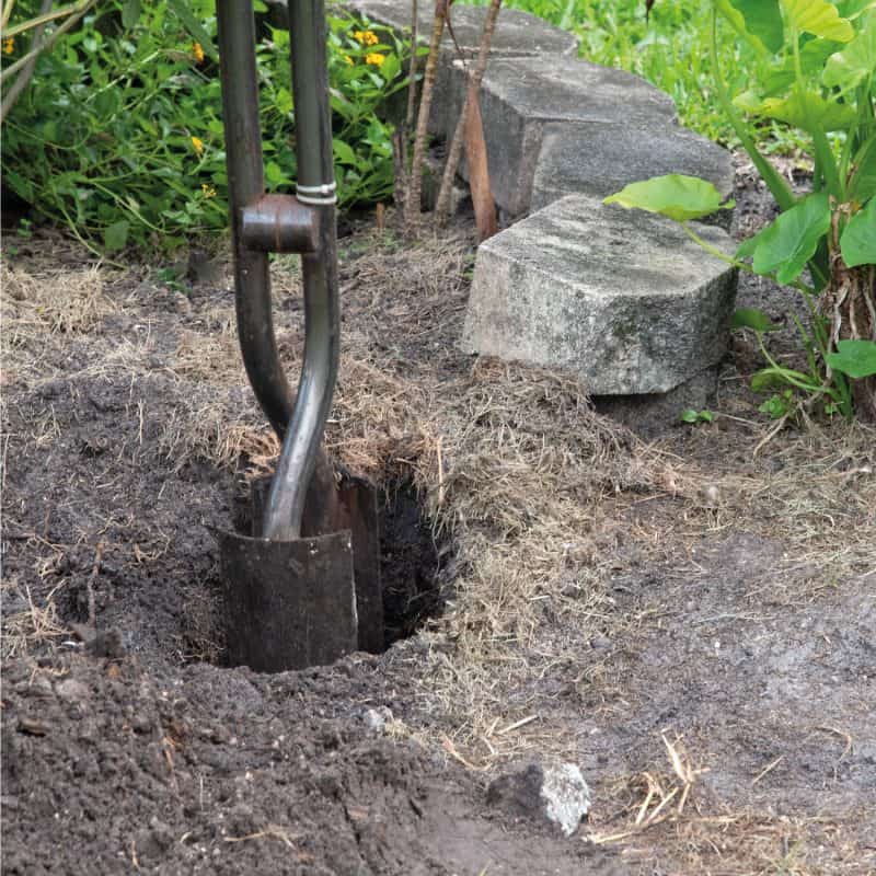 Steel-post-hole-digger-being-used-to-create-a-deep-hole-for-a-fence-post-with-concrete-bricks-and-green-plants-in-the-background-e1653384089381